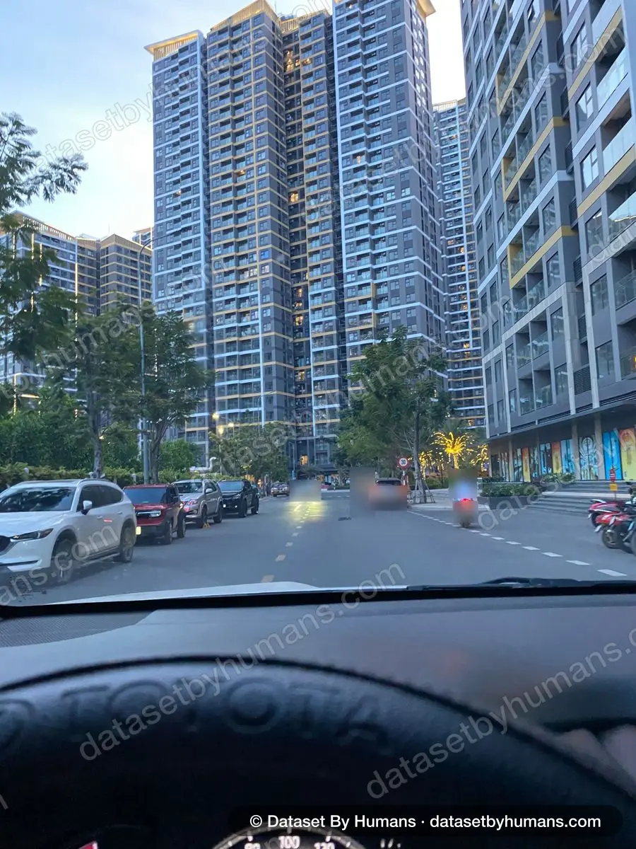 Street between high-rise apartment towers at dusk, seen from a car's driver seat, with parked cars and a passing motorbike