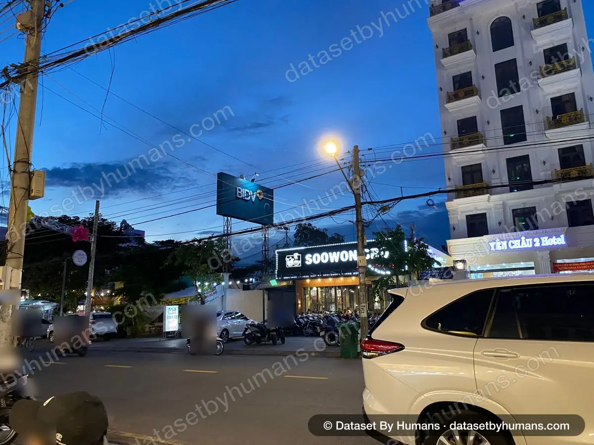 Evening street with motorbikes passing parked scooters outside shops and a hotel, under street lights and power lines