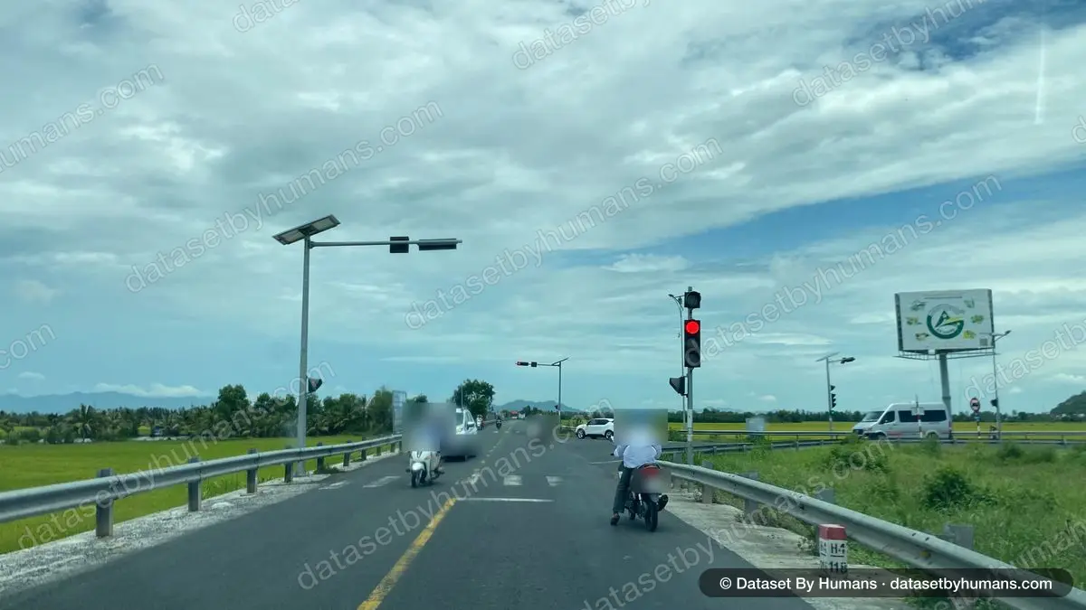 Two-lane rural road at a red traffic light with motorbikes and cars, rice fields on both sides under cloudy sky