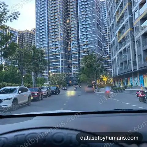 Street between high-rise apartment towers at dusk, seen from a car's driver seat, with parked cars and a passing motorbike