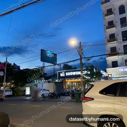 Evening street with motorbikes passing parked scooters outside shops and a hotel, under street lights and power lines