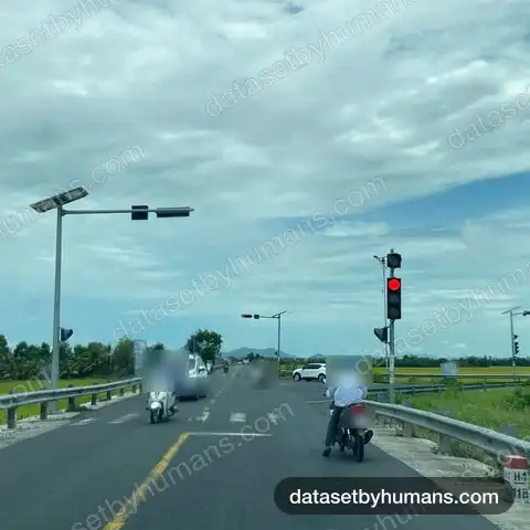 Two-lane rural road at a red traffic light with motorbikes and cars, rice fields on both sides under cloudy sky