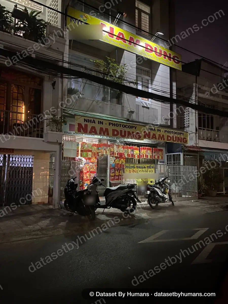 Night street view of a print and photocopy shop with large Vietnamese signs reading 'Nhà in - Photocopy Nam Dũng' and parked motorbikes