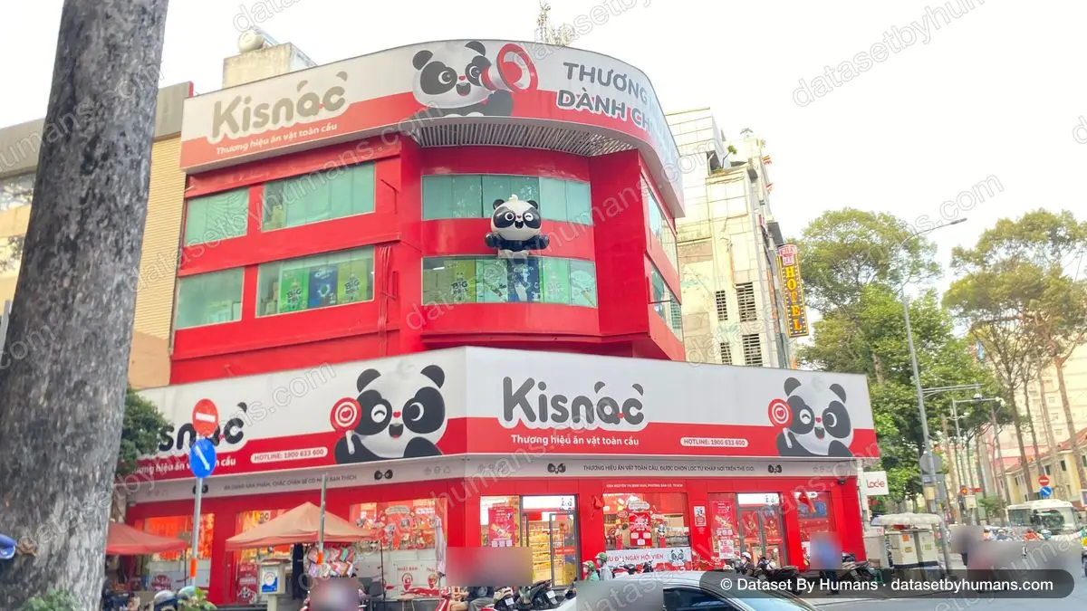 Corner snack store with large red and white Vietnamese signs, slogans and a hotline number, with street traffic below