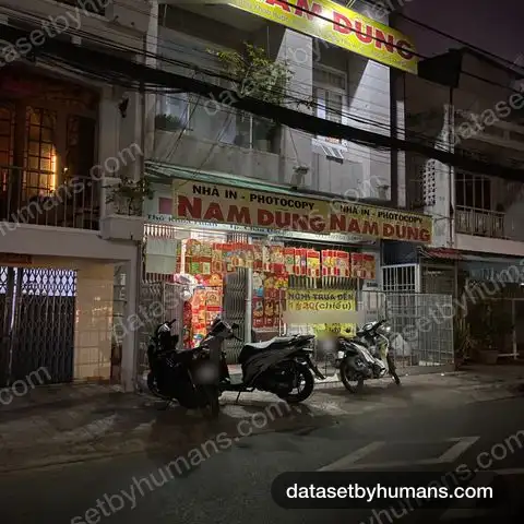Night street view of a print and photocopy shop with large Vietnamese signs reading 'Nhà in - Photocopy Nam Dũng' and parked motorbikes