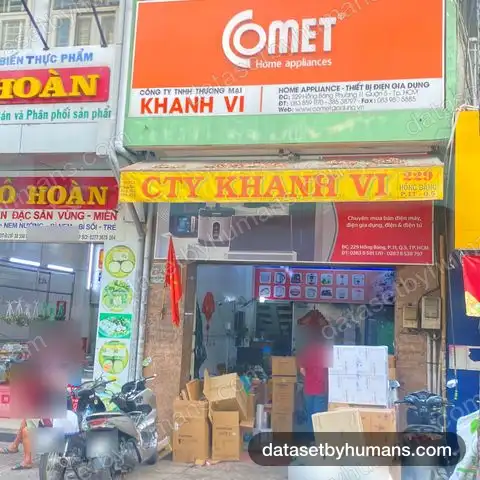 Shopfront of a home-appliance company with dense Vietnamese signage, neighbouring food shop signs and motorbikes on the pavement