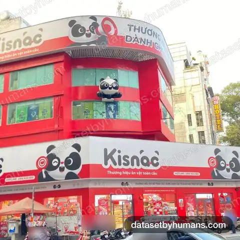 Corner snack store with large red and white Vietnamese signs, slogans and a hotline number, with street traffic below